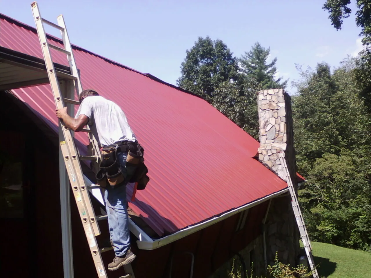 Roofer on a ladder beside a red metal roof and stone chimney — Soffit & Fascia Repair by Roofing Brothers in Lyndon